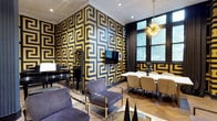 This single-floor space makes an immediate impression with its dramatic interior design, defined by bold black and gold Greek key-patterned wallcoverings that envelop nearly every wall, creating a cohesive and opulent atmosphere throughout. The space is thoughtfully arranged to serve multiple functions, seamlessly blending a lounge area, dining setting, and musical corner.

The lounge area features plush velvet armchairs with gold-toned metal frames, arranged around a glass-top coffee table. A wall-mounted flat-screen television is centrally positioned against the Greek key backdrop, adding a modern touch. A sleek black grand piano occupies one corner, accompanied by a full-length mirror, lending an air of sophistication and cultural refinement.

The dining area is anchored by a long marble-top table surrounded by elegant upholstered chairs, making it ideal for gatherings. A striking Sputnik-style brass chandelier hangs above, casting warm light across the setting. Nearby, large windows dressed with dark curtains allow natural light to filter in while offering views of greenery outside.

A classical black sculptural bust serves as a decorative centerpiece on the dining table, reinforcing the space's art-forward aesthetic. Herringbone-patterned wood flooring grounds the design, providing warmth beneath the bold décor. A decorative arched entryway with contrasting geometric wall art marks the transition to a vestibule area, completing this visually immersive environment.