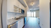 This thoughtfully designed studio offers a streamlined, open-concept layout that integrates the kitchen and living area into a single cohesive space. The architectural character is defined by striking exposed concrete ceilings, which lend an industrial-modern aesthetic throughout, complemented by warm hardwood-style flooring that runs continuously across the unit.

The kitchen is well-appointed with white flat-panel cabinetry extending floor to ceiling, providing generous storage. Stainless steel appliances — including a refrigerator, electric range, over-the-range microwave, and dishwasher — are paired with granite countertops and a classic white subway tile backsplash, creating a clean, contemporary finish. A deep single-basin sink with a modern pull-down faucet completes the workspace.

The living area is bright and airy, anchored by a large floor-to-ceiling window and glass door that opens to an outdoor balcony, flooding the room with natural light and offering expansive city views. A wall-mounted HVAC unit ensures year-round climate comfort.

The bathroom features granite countertops, white cabinetry, and hardwood-style flooring consistent with the rest of the unit, along with accessibility grab bars. Built-in shelving and storage units in the living area enhance the overall functionality of this efficiently planned space.