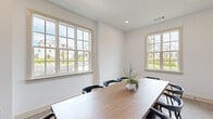 Located on Denali Drive in Mountain Park, Georgia, this single-room meeting space offers a clean, contemporary design well-suited for professional gatherings or collaborative work sessions. The room is defined by its crisp white walls and neutral-toned carpet, which together create a calm, focused atmosphere. A substantial natural wood conference table anchors the space, surrounded by dark, modern chairs that provide comfortable seating for a full group. Multiple large, multi-pane windows line the walls, flooding the interior with generous natural light and framing views of the surrounding neighborhood and greenery. Recessed ceiling lighting supplements the daylight, ensuring consistent illumination throughout. A standout architectural feature is the set of sleek, black-framed glass panel doors that open to an adjacent area, maintaining visual connectivity while defining the room's boundaries. Built-in closet doors with matte black hardware offer practical storage without interrupting the room's streamlined aesthetic. A framed landscape artwork adds a subtle decorative touch, enhancing the professional yet welcoming ambiance. The overall design balances functionality with refined simplicity, making this a polished and inviting space for meetings and presentations.