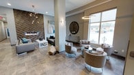 This sophisticated single-floor lounge space showcases a carefully curated contemporary design. A striking textured stone accent wall frames a sleek linear fireplace, serving as a dramatic focal point anchored by a stylish sofa and geometric coffee table. The space transitions seamlessly into a bright seating area bathed in natural light from large windows overlooking the exterior. An eclectic mix of upholstered barrel chairs, metallic side tables, and plush seating arrangements encourages conversation and relaxation throughout. Overhead, a sculptural sputnik chandelier and a circular halo pendant light add artistic flair, while wall sconces provide warm ambient illumination. Large-format porcelain tile flooring in a neutral tone grounds the space with a clean, polished look. An oversized decorative clock adorns the wall, adding a bold design statement. The overall palette of warm grays, taupe, and metallic accents creates a refined, modern ambiance that is both inviting and visually engaging.