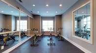 This single-floor fitness space is purpose-built as a cycling studio, designed to deliver a focused and motivating workout experience. The room is outfitted with multiple stationary spin bikes arranged across a durable rubber floor, which provides both cushioning and stability during high-intensity sessions. Large windows along the far wall invite natural light into the space, creating an open and energizing atmosphere. Recessed ceiling lights ensure consistent, even illumination throughout the room, complementing the natural light for use at any time of day. Full-length wall mirrors line one side of the studio, allowing participants to monitor their form and enhancing the sense of spaciousness within the room. The walls are finished in a cool, neutral gray tone with crisp white trim, contributing to a clean and professional aesthetic. A framed wall mirror on the adjacent side adds further depth to the space. The rubber flooring extends wall to wall, reinforcing the room's identity as a dedicated athletic environment. Overall, the studio combines practical, performance-oriented design with a polished interior finish, making it a well-equipped and visually cohesive space for cycling workouts.