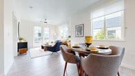 This thoughtfully designed single-floor residence offers a seamless blend of contemporary style and everyday functionality. The open-concept living and dining area is bathed in natural light, courtesy of large windows and glass French doors that lead to a private balcony overlooking a landscaped neighborhood. Light-toned hardwood-style flooring runs throughout the main living spaces, complementing the clean white walls and creating a bright, airy ambiance. A ceiling fan anchors the living room, which flows effortlessly into a dedicated dining area framed by additional windows. The kitchen features wood-tone cabinetry, subway tile backsplash, stainless steel appliances including a refrigerator and microwave, recessed lighting, and generous counter space — all centered around a versatile rolling kitchen island. The primary bedroom is a comfortable retreat with plush carpet, a walk-in closet with built-in shelving, and a window that fills the room with soft natural light. A second bedroom offers similar comfort and serene outdoor views. The bathroom is finished with floor-to-ceiling subway tile surrounding a deep soaking tub, accented by polished chrome fixtures. The private balcony extends the living space outdoors, offering open views of the surrounding community and greenery.