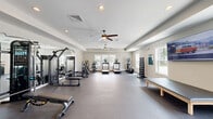 Located at 3021 Siskin Dr NW in Calabash, North Carolina, this fitness center offers a spacious, single-floor layout designed to accommodate a comprehensive workout experience. The open floor plan is finished with durable dark flooring suitable for high-traffic athletic use, while neutral-toned walls and a tray ceiling contribute to a clean, modern aesthetic throughout.

Natural light flows in through multiple windows positioned along the walls, creating a bright and inviting atmosphere. Two ceiling fans with integrated lighting provide consistent air circulation, complementing the recessed lighting overhead. A large wall-mounted flat-screen TV is positioned for easy viewing during workouts.

The cardio zone features multiple treadmills and elliptical machines, each equipped with individual display screens. A dedicated strength training area includes a multi-station cable machine, weight benches, and a full-length mirror wall that enhances the sense of space. A neatly organized dumbbell rack and a medicine ball storage unit round out the free weights section, offering a range of resistance options.

An additional padded workout bench provides space for stretching or recovery. Entry to the fitness center is provided through a glass-paneled door, adding a polished touch to the facility. The overall design balances functionality and comfort, making this fitness center a well-appointed amenity for residents of the Calabash community.