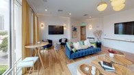 Located at 101–103 W Fortune St in the heart of Tampa, FL, this single-floor amenity lounge offers a thoughtfully designed communal space that blends coastal charm with contemporary sophistication. The open-plan layout is unified by warm hardwood flooring and bright white walls, creating an inviting atmosphere throughout.

The space is anchored by multiple seating arrangements featuring deep navy tufted sofas, velvet accent chairs, and an eclectic mix of sculptural wood coffee tables and gold-framed wire chairs. Statement pendant lighting — including clusters of brushed brass globe fixtures — adds warmth and visual interest overhead. Bold artwork and a decorative oar wall installation reinforce the coastal narrative, complemented by a striking blue-and-white striped accent wall.

A built-in wet bar area with white cabinetry, a butcher block counter, a wine refrigerator, and a sink provides a functional entertaining hub. Recycling and waste stations near the entrance support everyday usability.

Floor-to-ceiling windows along one wall flood the space with abundant natural light and frame sweeping views of the Tampa skyline, lush greenery, and palm tree-lined streets below. Gold drapery panels frame the windows, adding a refined finishing touch. The overall ambiance is vibrant yet relaxed — ideal for socializing and community gatherings.