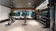 Located at 100 Callan Ave in San Leandro, CA, this well-appointed fitness center offers a thoughtfully designed, single-floor workout facility with a generous open floor plan. The space features a striking contemporary aesthetic, defined by dark ceilings with sleek surface-mounted cylindrical lighting, polished concrete floors, and warm wood-toned accent flooring in the cardio zone.

The main workout area is divided into distinct functional zones. The cardio section houses a row of treadmills, elliptical trainers, and interactive cycling bikes, each equipped with digital screens, set atop warm hardwood flooring and backed by large mirrors and wall-mounted televisions. A dedicated free weights area features fully stocked dumbbell and kettlebell racks alongside adjustable benches, framed by a distinctive arched wood-paneled mirror wall that adds depth and architectural character.

Floor-to-ceiling windows along one wall flood the space with natural light and provide views of the surrounding greenery outdoors. The welcoming entry area features a built-in wooden cubby console, tasteful artwork, and potted plants that lend a refined, boutique atmosphere. Structural columns finished in deep charcoal add an industrial touch, while the mirrored walls throughout visually expand the space. The overall ambiance is clean, modern, and motivating.