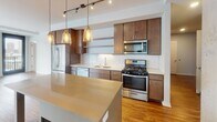 Located at 243 Chicago Ave S in Minneapolis, this single-floor unit offers a well-designed layout with modern finishes and notable urban views. The open-concept kitchen is a central highlight, featuring rich wood cabinetry, a white subway tile backsplash, stainless steel appliances including a gas range, microwave, dishwasher, and French door refrigerator, and a light-toned island beneath a cluster of Edison-style pendant lights. Track lighting overhead complements the warm hardwood-style flooring that flows through the main living areas. A sliding barn door adds a stylish architectural detail connecting spaces. The living area is bright and spacious, anchored by large floor-to-ceiling windows that frame sweeping Minneapolis skyline views, with exposed concrete columns adding an industrial character. The bedroom continues the city views through a generous window and features plush carpeting. Two bathrooms are finished with white subway tile; one includes a glass-enclosed walk-in shower with chrome fixtures and a floating wood vanity, while the other offers a full soaking tub with tiled surround and gray tile flooring. In-unit laundry is also present, adding everyday convenience to this contemporary urban residence.