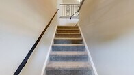 Located at 1101 Ferry Plantation Rd in Virginia Beach, Virginia, this well-laid-out three-bedroom, two-bathroom home offers a comfortable and functional floor plan across a single level of living space. The open living area is finished with neutral-toned carpet and light-colored walls, creating a bright and welcoming atmosphere. French-style doors connect the living space to the kitchen, which features white shaker cabinetry, light wood-look flooring, stainless steel appliances including a refrigerator and electric range, and a utility room conveniently accessible nearby. Each of the three bedrooms is carpeted and benefits from natural light through windows with outdoor views. The spacious primary bedroom includes a walk-in closet, while additional bedrooms are served by standard closets with bifold doors. The two bathrooms are finished with mosaic tile flooring; one features a full tub-shower combination with a grab bar, and the other includes a white vanity with under-cabinet storage and a well-lit mirror. A hallway with decorative wrought-iron stair railings and overhead lighting connects the main areas of the home. The overall aesthetic is clean, neutral, and move-in ready.