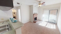 This single-story residence offers a well-organized and functional layout across several comfortable rooms. The living area serves as the heart of the home, featuring plush carpeted flooring, a ceiling fan with light fixture, and a classic brick fireplace that adds warmth and character to the space. Large vertical-blind-covered sliding doors allow generous natural light to pour in, creating a bright and inviting atmosphere. The living area connects seamlessly to the kitchen, which is equipped with dark espresso-toned cabinetry, light countertops, black appliances including a range and refrigerator, and wood-look flooring that grounds the space with a modern contrast. A breakfast bar counter separates the kitchen from the living area, adding convenience and an open feel. The bedroom is generously sized with soft carpeting, a ceiling fan, and a window with blinds that lets in plenty of natural light, offering a calm and restful environment. The bathroom features a wide vanity with dark cabinetry, a large mirror with overhead lighting, and a separate tub and shower area. A dedicated laundry space with a full-size washer and dryer adds practicality. Throughout the home, a neutral color palette and textured ceilings provide a clean, cohesive aesthetic.