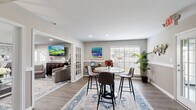Located at 2500 E James Ave in Baytown, TX, this thoughtfully designed single-floor space offers a cohesive and welcoming environment across three distinct areas. The living room serves as the central gathering space, featuring recessed lighting, a large wall-mounted TV, a bold navy blue sofa, paired gray accent chairs, and a glass-top coffee table set atop a patterned area rug. A striking stacked stone accent wall with a built-in bar counter and bar seating adds a sophisticated touch to one end of the living area. Adjacent to the living room, the dining area is bright and airy, benefiting from large windows with horizontal blinds that allow ample natural light. A round high-top table with modern bar-height chairs occupies the center, complemented by a wooden console table and vibrant artwork. French glass doors connect the dining room to the living space, enhancing the open flow between rooms. The office features a clean, functional layout with a half-wall glass partition, neutral tones, and wood-look flooring that runs consistently throughout the space. A wide hallway with framed artwork and a classic wall clock provides a polished transition between areas. The overall aesthetic is modern and inviting, with warm neutral walls, consistent hardwood-style flooring, and carefully curated décor.