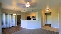 This single-floor residence offers an open and functional layout designed for comfortable everyday living. At the heart of the home is a well-appointed kitchen featuring white cabinetry, black appliances including a refrigerator, range, and microwave, all anchored by a wraparound peninsula that provides additional counter space and serves as a natural divider between the kitchen and the surrounding living areas. A ceiling fan with a light fixture is centrally positioned overhead, keeping the space bright and well-ventilated. The open-concept design allows the kitchen to flow seamlessly into the adjoining dining and living areas, creating a connected, sociable atmosphere. The flooring transitions from warm hardwood-style planks in the main living area to plush carpet in the lounge space, adding texture and comfort underfoot. A pendant light fixture adds a decorative touch near the dining area. Hallways branch off toward what appear to be private rooms and additional spaces, including a tiled area visible in the background, suggesting a bathroom or utility room nearby. The neutral wall tones and consistent lighting throughout contribute to a clean, inviting ambiance that is both practical and welcoming.
