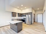 This thoughtfully designed single-floor unit offers a clean, modern aesthetic throughout. The kitchen serves as the heart of the space, showcasing dark espresso cabinetry with sleek hardware, light granite-style countertops, and a full suite of stainless steel appliances including a refrigerator, range, microwave, and dishwasher. A movable kitchen island with a white countertop provides additional prep and storage space, adding versatility to the layout. Light-toned hardwood-style flooring runs throughout, contributing to a bright and cohesive look. Recessed lighting in the kitchen ensures excellent visibility, while the adjacent living area benefits from impressively tall ceilings that amplify the sense of openness. A large window, paired with a transom window above, floods the living area with natural light and offers views of surrounding greenery. The hallway connects the main living areas to the well-appointed bathroom, which features a wide quartz vanity countertop, an undermount sink, dark cabinetry, and a decorative tile backsplash accent. The overall ambiance is contemporary and functional, with consistent finishes that create a polished, move-in-ready environment.