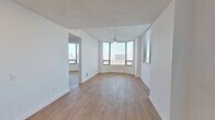 This single-floor unit offers a clean, contemporary design characterized by crisp white walls, light wood-tone flooring, and an abundance of natural light. The living area is a standout feature, anchored by a curved bay window that wraps around one corner of the room, flooding the space with daylight and framing elevated views of the surrounding cityscape. A ceiling fan with a pendant-style light fixture serves the area, and an adjacent room—accessible through an open doorway—provides additional flexible living space with its own windows and city views.

The layout, visible in the floor plan model, reveals a well-organized arrangement of living, sleeping, and service areas. The kitchen is fitted with white cabinetry, light stone-look countertops, a stainless steel electric range, and an over-the-range microwave, offering a functional and tidy cooking environment.

The bedroom features light wood flooring, a ceiling fan, windows on two walls providing cross-ventilation and views, and a wide sliding-door closet for ample storage. The bathroom is finished in a modern monochromatic palette, with vertically stacked white subway tile surrounding the tub-shower combination, matte black fixtures and hardware, a white vanity with stone countertop, and wood-tone flooring that ties the space together cohesively.
