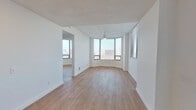 This single-floor unit offers a clean, contemporary design characterized by crisp white walls, light wood-tone flooring, and an abundance of natural light. The living area is a standout feature, anchored by a curved bay window that wraps around one corner of the room, flooding the space with daylight and framing elevated views of the surrounding cityscape. A ceiling fan with a pendant-style light fixture serves the area, and an adjacent room—accessible through an open doorway—provides additional flexible living space with its own windows and city views.

The layout, visible in the floor plan model, reveals a well-organized arrangement of living, sleeping, and service areas. The kitchen is fitted with white cabinetry, light stone-look countertops, a stainless steel electric range, and an over-the-range microwave, offering a functional and tidy cooking environment.

The bedroom features light wood flooring, a ceiling fan, windows on two walls providing cross-ventilation and views, and a wide sliding-door closet for ample storage. The bathroom is finished in a modern monochromatic palette, with vertically stacked white subway tile surrounding the tub-shower combination, matte black fixtures and hardware, a white vanity with stone countertop, and wood-tone flooring that ties the space together cohesively.