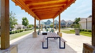 Located at 760 W Bedford Euless Rd in Hurst, Texas, this thoughtfully designed outdoor community space serves as a central gathering hub within a well-maintained residential neighborhood. The focal point of the space is a substantial wood-beam pergola supported by stone-base columns, providing welcome shade over a generous concrete patio. The pergola's warm natural timber construction creates an inviting, open-air atmosphere ideal for relaxation and social gatherings.

The patio area is furnished with Adirondack chairs and additional outdoor seating, offering comfortable spots to enjoy the Texas sunshine and fresh air. Concrete walkways connect the pergola area to surrounding pathways, ensuring easy accessibility throughout the space. A secondary seating area nearby features lounge chairs, expanding the capacity for residents to gather.

The grounds are beautifully landscaped with neatly trimmed shrubs, young trees, and lush green lawns that provide a clean, park-like setting. Black iron fencing borders portions of the space, adding a classic aesthetic while defining the area. Views from the pergola look out across the broader neighborhood streetscape, where a variety of craftsman and traditional-style homes with pitched rooflines create a charming residential backdrop. The combination of mature plantings, open lawn areas, and quality outdoor amenities makes this space a welcoming extension of the community.