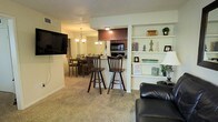 This single-floor residence is designed with an open-concept layout that integrates the living, dining, and kitchen areas into one cohesive space. The living area features plush carpeting underfoot and is anchored by a wall-mounted flat-screen TV, alongside built-in shelving that provides both display and storage space — a standout architectural detail that adds character to the room. A comfortable leather sofa completes the seating arrangement.

A raised breakfast bar with bar-height seating for two serves as a natural divider between the living space and the kitchen, offering a casual dining option while maintaining the open feel of the layout. The kitchen, positioned at the rear, is equipped with dark cabinetry, black appliances including a built-in microwave and oven, and is illuminated by recessed lighting.

Adjacent to the kitchen, a dedicated dining area comfortably accommodates a full dining table and chairs, lit by a chandelier overhead. The warm, neutral color palette used throughout — including the beige carpet and white walls — gives the space a cohesive and welcoming ambiance suitable for everyday living and entertaining.