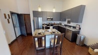 Located along the I-35 E Frontage Road in Denton, TX, this single-floor residence offers a contemporary design with an open, airy layout. The kitchen is a standout feature, boasting soaring ceilings that amplify the sense of space, sleek gray upper cabinetry paired with dark lower cabinets, and granite countertops extending across a central island with bar seating. Stainless steel appliances — including a refrigerator, range, microwave, and dishwasher — complete the space, while pendant lighting adds a refined touch. Rich hardwood-style flooring flows throughout the common areas, grounding the modern aesthetic. The living area is an intimate yet functional space, benefiting from natural light streaming through a large window with exterior door access, complemented by high walls that lend an open character. The bedroom is a calm retreat, featuring plush carpeting, a large window with outdoor views, and direct access to a walk-in closet offering generous storage. The bathroom is well-appointed with granite vanity, dark cabinetry, and a full tiled tub-shower surround. Throughout the residence, a cohesive palette of grays, dark wood tones, and neutral whites creates a polished, modern ambiance suited to comfortable everyday living.