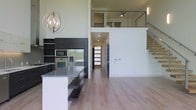 This contemporary two-story space is defined by its striking open-concept design and impressive double-height ceilings that create a sense of expansive verticality. The main level centers around a well-appointed kitchen finished in dark cabinetry with a gray countertop island that offers additional seating. Full-height cabinetry provides ample storage, while stainless steel appliances — including a built-in oven, microwave, and refrigerator — complete the functional and stylish kitchen setup. An elegant orb-style chandelier hangs overhead, adding a refined focal point to the space. Light-toned hardwood flooring runs throughout the main level, contributing warmth and continuity to the open floor plan. A hallway leads toward a modern front entry door with frosted glass panels, allowing natural light to filter through. The upper mezzanine level overlooks the main living area through sleek cable-rail balustrades, offering an airy, connected feel between floors. A modern staircase with open risers and cable railing provides access to the upper level, which features a ceiling fan and additional living or sleeping space. The overall ambiance is clean, minimalist, and modern throughout.