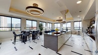 This elegantly designed single-floor space showcases a seamless open-concept layout that integrates a gourmet kitchen, dining area, and lounge into one cohesive, light-filled environment. The kitchen serves as a centerpiece, featuring a large island with a dark stone countertop, a mosaic tile backsplash, stainless steel appliances, and a statement range hood — all complemented by high-gloss cabinetry. Tray ceilings with warm gold accents frame each zone, and sculptural ring chandelier lighting adds a dramatic, contemporary touch throughout. The dining area is appointed with a sleek black high-top table and leather bar-style seating, positioned beneath large black-framed windows that flood the space with natural light and sweeping city skyline views. The lounge area features a striking curved blue velvet sofa, a round marble-topped coffee table, and layered textures that create an inviting yet upscale atmosphere. Polished light stone tile flooring with bold dark inlay accents runs throughout, tying each zone together with a refined, modern aesthetic. Sliding glass doors lead to an outdoor terrace, extending the living space and offering direct access to open-air city views — making this an exceptional space for both everyday living and entertaining.