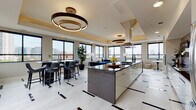 This elegantly designed single-floor space showcases a seamless open-concept layout that integrates a gourmet kitchen, dining area, and lounge into one cohesive, light-filled environment. The kitchen serves as a centerpiece, featuring a large island with a dark stone countertop, a mosaic tile backsplash, stainless steel appliances, and a statement range hood — all complemented by high-gloss cabinetry. Tray ceilings with warm gold accents frame each zone, and sculptural ring chandelier lighting adds a dramatic, contemporary touch throughout. The dining area is appointed with a sleek black high-top table and leather bar-style seating, positioned beneath large black-framed windows that flood the space with natural light and sweeping city skyline views. The lounge area features a striking curved blue velvet sofa, a round marble-topped coffee table, and layered textures that create an inviting yet upscale atmosphere. Polished light stone tile flooring with bold dark inlay accents runs throughout, tying each zone together with a refined, modern aesthetic. Sliding glass doors lead to an outdoor terrace, extending the living space and offering direct access to open-air city views — making this an exceptional space for both everyday living and entertaining.