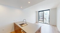 This thoughtfully designed one-bedroom, one-bathroom residence offers a clean, modern aesthetic with an efficient and functional layout. Light hardwood flooring flows seamlessly throughout the entire unit, creating a warm and cohesive feel. The combined kitchen and living area serves as the heart of the home, anchored by a generous kitchen island with a quartz-style countertop, undermount sink, and warm wood cabinetry. Stainless steel appliances — including a refrigerator, oven, microwave, and dishwasher — complete the well-appointed kitchen, which also features a mosaic tile backsplash. Floor-to-ceiling windows in the living area and bedroom flood the space with natural light and frame striking elevated views of the surrounding cityscape. The bedroom is a calm, well-proportioned retreat with large windows that continue the urban panorama. A dedicated closet provides practical storage. The bathroom is finished with large-format neutral tile surrounding a full soaking tub with shower, complemented by sleek chrome fixtures and wood-toned vanity details. Recessed lighting and crisp white walls throughout give the unit a bright, contemporary feel well-suited for modern urban living.
