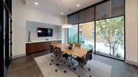 This single-floor facility combines a dedicated computer lab and a professional conference room, thoughtfully designed for both individual productivity and collaborative meetings. The computer lab features a marble-topped workstation built into a warm wood-paneled alcove, accommodating multiple workstations equipped with desktop computers and task lighting. Large-format floor tiles in a cool gray tone anchor the space and flow seamlessly throughout both areas. The conference room is bright and open, anchored by a solid wood rectangular table surrounded by ergonomic rolling chairs. A circular pendant light fixture hangs overhead, complementing the recessed lighting above. One wall showcases a built-in wood credenza topped with a wall-mounted flat-screen display set against textured wallcovering, providing a polished focal point. Floor-to-ceiling steel-framed glass walls flood the conference area with natural light and offer unobstructed views of a lush outdoor courtyard with mature trees and landscaped greenery. Original artwork adorns the walls, adding a refined, creative touch. The overall ambiance is contemporary and professional, balancing clean architectural lines with warm wood accents and an inviting connection to the outdoors.