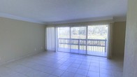 This single-floor space is defined by its bright, open living area that draws the eye immediately toward a expansive wall of sliding glass doors. The doors span nearly the full width of the room, flooding the interior with natural light and providing direct access to an outdoor balcony. Vertical blinds offer adjustable light control while maintaining the connection to the outdoors.

The living area is finished with light-toned ceramic tile flooring laid in a classic grid pattern, contributing to a clean and airy aesthetic. The ceiling features a textured finish and is accented by crown molding where it meets the walls, adding a subtle architectural detail to the otherwise understated interior.

Beyond the balcony's railing, a lush, wooded landscape stretches into the distance, providing a tranquil natural backdrop that enhances the sense of openness and calm within the space. The neutral wall tones complement the natural light, creating a versatile interior environment. Overall, the space offers a seamless connection between indoor living and the surrounding natural scenery.