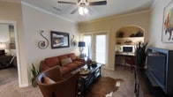 This single-floor space exudes a warm and inviting ambiance, thoughtfully designed with a cohesive Southwestern aesthetic. The main living area centers around rich cognac leather seating arranged over a cowhide area rug, creating an intimate and comfortable gathering space. A dark-framed coffee table serves as a functional focal point, while decorative wrought-iron wall accents and framed artwork add character to the neutral-toned walls. Crown molding lends an elegant architectural detail, and a ceiling fan with a light fixture provides both comfort and illumination. A sliding glass door dressed with plantation shutters allows natural light to filter into the room while offering privacy. One of the standout features of this space is the built-in arched nook tucked along one wall, outfitted with open shelving for display and a dedicated desk area — ideal for a home office setup. The shelving showcases decorative pieces that complement the room's earthy tones. Carpeted flooring throughout adds to the cozy feel of the space. Overall, the layout balances functional living and workspace needs within a warm, character-rich environment that feels both purposeful and welcoming.
