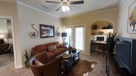 This single-floor space exudes a warm and inviting ambiance, thoughtfully designed with a cohesive Southwestern aesthetic. The main living area centers around rich cognac leather seating arranged over a cowhide area rug, creating an intimate and comfortable gathering space. A dark-framed coffee table serves as a functional focal point, while decorative wrought-iron wall accents and framed artwork add character to the neutral-toned walls. Crown molding lends an elegant architectural detail, and a ceiling fan with a light fixture provides both comfort and illumination. A sliding glass door dressed with plantation shutters allows natural light to filter into the room while offering privacy. One of the standout features of this space is the built-in arched nook tucked along one wall, outfitted with open shelving for display and a dedicated desk area — ideal for a home office setup. The shelving showcases decorative pieces that complement the room's earthy tones. Carpeted flooring throughout adds to the cozy feel of the space. Overall, the layout balances functional living and workspace needs within a warm, character-rich environment that feels both purposeful and welcoming.