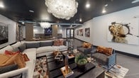 This expansive single-floor amenity space blends contemporary industrial design with upscale residential comfort across several distinct zones. The main lounge area makes an immediate impression with its dark, painted ceiling, exposed ductwork, and dramatic globe chandeliers casting warm, ambient light over large-format tile flooring. Generous seating arrangements—featuring patterned accent chairs, neutral sofas, and bold throw pillows in burnt orange and earth tones—create an inviting gathering atmosphere. Structural columns clad in warm wood tones anchor the open layout while curated artwork displayed throughout reinforces an urban, gallery-like aesthetic.

The full kitchen is a standout feature, highlighted by a curved granite-topped island with gold metallic detailing, sleek acrylic barstools, stainless steel appliances, white cabinetry, and a striking herringbone tile backsplash. Pendant lighting and views to the outdoor area enhance the space's functionality and style.

The game room offers a billiards table, ping pong table, wall-mounted television, and direct access to an outdoor patio through large sliding glass doors, seamlessly connecting interior and exterior social spaces. An entry corridor with large-format tile and bold framed artwork provides a polished transition throughout the space.
