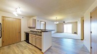 This single-floor residence showcases an open-concept layout that seamlessly connects the kitchen and living areas, creating a spacious and functional environment. The kitchen features a peninsula-style counter with dark countertops, light wood cabinetry, a double-basin sink, and a dishwasher. The peninsula serves as both a workspace and a natural divider between the kitchen and the adjoining living area, enhancing the sense of flow throughout the space.

The flooring transitions from light hardwood-style planks in the kitchen area to carpeting in the living space, adding texture and visual distinction between the two zones. The living area is generously sized and benefits from natural light streaming through a sliding glass door, which also provides access to an outdoor area. A ceiling fan with a light fixture is centrally positioned in the living room, complemented by a chandelier near the dining area, offering layered lighting options.

The textured ceilings add a classic touch throughout, while neutral wall tones keep the space feeling bright and versatile. Overall, the open layout and functional design make this a practical and comfortable living space.