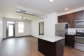 This single-floor unit offers a well-designed, open-concept layout that seamlessly connects the kitchen and living area. The kitchen is a standout feature, outfitted with rich dark-wood cabinetry, light quartz countertops, a full suite of stainless steel appliances including a refrigerator, electric range, microwave, and dishwasher, as well as a central island that provides additional prep and seating space. Open wall-mounted shelving adds a modern, industrial touch alongside the exposed metallic ductwork that runs across the ceiling, lending the space an urban loft aesthetic. Hardwood-style flooring flows throughout the entire unit, providing a warm, cohesive look. The living area is generously sized, illuminated by recessed lighting and a ceiling fan, with a main entry door flanked by windows that allow natural light to fill the space. The bedroom is comfortably proportioned, featuring its own ceiling fan and a window with an exterior view. A hallway connects the bedroom to the bathroom, which includes a vanity with shelving. A dedicated laundry closet houses a stacked Whirlpool washer and dryer, adding everyday convenience to this thoughtfully appointed unit.