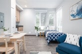 This single-floor studio unit offers a thoughtfully organized layout that maximizes every square foot of available space. The main living area combines sleeping, lounging, and dining functions in one open room, featuring light wood-tone flooring, neutral grey walls, and recessed lighting throughout. Large windows fitted with horizontal blinds line the exterior wall, allowing an abundance of natural light to fill the space while offering views of surrounding greenery. The sleeping area accommodates a twin bed and a comfortable accent chair, creating a cozy yet functional arrangement. A compact dining table with seating is positioned near the entry, complemented by a low open-shelf console for additional storage. The kitchenette is tucked into an alcove and is equipped with warm wood cabinetry, a dark countertop, a single-basin sink, a microwave, and a compact refrigerator — providing all the essentials for everyday meal preparation. The bathroom features a clean, modern design with a white vanity sink, a framed mirror, and a shower enclosed by a decorative white curtain. A solid dark entry door with a secure latch completes the unit. Overall, the space maintains a bright, clean, and modern aesthetic well-suited for comfortable single-occupancy living.