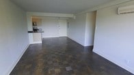 This single-floor space showcases a thoughtfully designed open-concept layout that seamlessly connects the kitchen and living areas into one cohesive, functional environment. The interior is characterized by rich, dark parquet flooring arranged in a classic block pattern, adding warmth and visual texture throughout the main living space.

The kitchen is positioned toward one end of the open floor plan and features light wood cabinetry, dark countertops, and stainless steel appliances, including a microwave. A peninsula-style counter provides a natural transition between the kitchen and the living area, offering both additional workspace and casual seating potential.

The living area is generously proportioned, offering ample room for a variety of furniture arrangements. Crisp white walls and clean white baseboards contribute to a bright, neutral palette that enhances the sense of openness. A structural beam element is visible overhead, adding an architectural detail to the ceiling plane. A white interior door provides access to additional areas of the space.

Overall, the space presents a clean, modern aesthetic with a practical layout, making it well-suited for everyday living and entertaining alike.