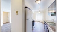 Located at 1646 W Lorain St in Monroe, Michigan, this single-floor, one-bedroom, one-bathroom unit offers a practical and comfortable layout with distinct living spaces. The galley-style kitchen is well-appointed with dark wood cabinetry, granite-style countertops, a double-basin sink, an electric range, a full-size refrigerator, and a range hood. Warm wood-look flooring runs throughout the kitchen, which also benefits from natural light streaming in through a vertical-blind-covered window at the far end. A dedicated storage or pantry area is accessible directly from the kitchen. The living area is generously sized, featuring plush carpeting, multiple windows with vertical blinds that allow for abundant natural light, and a wall-mounted air conditioning unit alongside baseboard heating for year-round comfort. The bedroom is a well-proportioned, carpeted room with a ceiling light fixture and its own entry door. The full bathroom includes a tub and shower combination, a toilet, a single vanity with dark wood cabinetry, and wood-look tile flooring. The overall design is clean and neutral throughout, with white walls and simple finishes that create a bright, functional living environment.