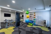 This single-floor fitness center is a comprehensively outfitted facility designed to support a wide range of workout routines. The space is anchored by a bold geometric carpet in charcoal, grey, and yellow tones that gives the room a modern, energetic aesthetic throughout.

The cardio zone features multiple treadmills and elliptical machines, each equipped with individual screens for entertainment during workouts. A dedicated strength training area includes a cable machine, a weight bench, and a rack of dumbbells, complemented by large wall-mounted mirrors that enhance the sense of openness. A sleek, freestanding Forma fitness pod provides an additional interactive training option.

A well-stocked functional training station offers stability balls in various sizes, resistance bands, medicine balls, foam rollers, and exercise mats — providing ample options for flexibility, balance, and core training. Wall-mounted televisions are positioned throughout to keep users engaged.

A welcoming amenity station near the entrance features infused water dispensers, towels, and refreshment supplies. The fitness center operates 24 hours a day and is reserved for residents. A clean, tiled restroom is conveniently accessible directly from the workout floor. Recessed lighting throughout ensures bright, even illumination across the entire space, creating a focused and motivating environment for exercise.