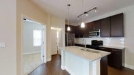 This single-floor space showcases a thoughtfully designed open-concept layout that seamlessly connects the kitchen and living areas. The kitchen is a standout feature, boasting rich dark cabinetry that contrasts beautifully with the light granite countertop island, which also functions as a breakfast bar with an undermount sink. Stainless steel appliances — including a refrigerator, built-in microwave, and electric range — complement the modern aesthetic. Pendant lighting hangs elegantly above the island, while a track lighting system provides additional illumination over the cooking area. The warm hardwood flooring flows throughout the main living space, adding a welcoming sense of continuity. Neutral, light-toned walls keep the interior bright and versatile. Adjacent to the kitchen, a hallway leads to additional rooms, including a bathroom with natural light filtering through a window, and a carpeted bedroom area that adds a softer texture to the overall design. The layout is efficient and well-organized, making excellent use of the available space. Overall, the unit strikes a clean, contemporary feel with a warm residential ambiance suitable for comfortable everyday living.