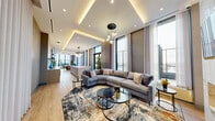 Located at 500 Upper Wellington St in Hamilton, Ontario, this elegantly designed single-floor space offers a seamlessly connected open-concept layout integrating a living room, dining room, and kitchen. The interior is characterized by soaring floor-to-ceiling windows that flood the space with natural light and provide expansive outdoor views, complemented by sheer draping curtains that add softness to the contemporary aesthetic.

The living area is anchored by a striking marble-clad fireplace wall, above which a wall-mounted television is integrated, creating a refined focal point. A generous sectional sofa, layered area rugs, and glass-topped circular coffee tables contribute to the lounge's inviting atmosphere. Statement globe pendant lighting adds an artistic dimension overhead.

The kitchen is a standout feature, showcasing a long, waterfall-edge quartz island with bar seating, warm wood-toned cabinetry, white quartz countertops, and a sculptural multi-globe chandelier. Recessed cove lighting above the cabinetry enhances the polished ambiance.

The dining area accommodates a large rectangular table surrounded by upholstered chairs, bathed in natural light from adjacent floor-to-ceiling windows. Light hardwood flooring flows uniformly throughout, unifying the open, contemporary design with a warm, welcoming character.