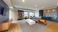 Located at 410 W Lake Street in Minneapolis, this well-appointed amenity space offers a generously sized, single-floor layout designed for gathering and relaxation. The interior living room features warm light hardwood flooring, recessed lighting, and large windows that frame elevated views of the surrounding Minneapolis cityscape, filling the space with abundant natural light. A bold, colorful mural-style artwork anchors one wall, adding a vibrant artistic focal point. Seating areas are arranged to encourage both casual conversation and comfortable lounging, complemented by a dark-brick accent wall with a wall-mounted television and a mid-century-style wood media console. A kitchenette with dark cabinetry and a sink provides convenient service support for events and gatherings. The interior flows seamlessly to an expansive rooftop terrace through floor-to-ceiling glass doors. Outside, a pergola-covered section shelters a built-in grill station and outdoor dining table, while the open-air portion features a central fire pit surrounded by mixed seating — including benches, lounge chairs, and accent stools — strung with overhead bistro lights. Panoramic views of the Minneapolis skyline make this rooftop terrace a standout feature of the space.