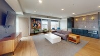 Located at 410 W Lake Street in Minneapolis, this well-appointed amenity space offers a generously sized, single-floor layout designed for gathering and relaxation. The interior living room features warm light hardwood flooring, recessed lighting, and large windows that frame elevated views of the surrounding Minneapolis cityscape, filling the space with abundant natural light. A bold, colorful mural-style artwork anchors one wall, adding a vibrant artistic focal point. Seating areas are arranged to encourage both casual conversation and comfortable lounging, complemented by a dark-brick accent wall with a wall-mounted television and a mid-century-style wood media console. A kitchenette with dark cabinetry and a sink provides convenient service support for events and gatherings. The interior flows seamlessly to an expansive rooftop terrace through floor-to-ceiling glass doors. Outside, a pergola-covered section shelters a built-in grill station and outdoor dining table, while the open-air portion features a central fire pit surrounded by mixed seating — including benches, lounge chairs, and accent stools — strung with overhead bistro lights. Panoramic views of the Minneapolis skyline make this rooftop terrace a standout feature of the space.