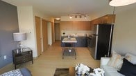 This single-floor space is designed with a modern, open-concept layout that seamlessly connects the kitchen and living areas. The kitchen is a standout feature, outfitted with warm wood upper cabinetry, sleek dark lower cabinets, and stainless steel appliances including a full-size refrigerator and a stove with range hood. A freestanding island with a light countertop provides additional prep space and a casual gathering spot. Light-toned laminate flooring runs throughout, lending a clean and cohesive look to the space. Track lighting overhead in the kitchen area ensures ample illumination for cooking and entertaining. A hallway off the main living area leads to additional rooms, including what appears to be a bedroom and storage or closet space, each accessed through clean, simple door designs consistent with the unit's contemporary aesthetic. The open living area, visible in the foreground, offers a comfortable and functional space for relaxation. Overall, the space presents a well-appointed, efficient layout with modern finishes that create a bright and welcoming atmosphere throughout.