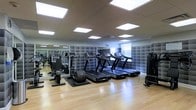 This fitness center offers a clean, modern environment designed to support a full range of exercise routines. The space features warm hardwood flooring throughout, providing a polished and functional surface well-suited to both cardio and strength training activities. The ceiling is fitted with a grid of recessed panel lighting that ensures the entire room is evenly and brightly illuminated, creating an energizing atmosphere. Natural light supplements the artificial lighting through windows along one wall, adding a sense of openness to the space. A row of treadmills equipped with individual digital display screens lines the back wall, offering users an interactive cardio experience. Additional cardio equipment, including stationary bikes and elliptical machines, is positioned throughout the room to maximize the use of available floor space. The back wall features a distinctive padded tile design in neutral grey tones, contributing both an aesthetic accent and acoustic functionality. A large mirror is mounted along one wall, enhancing the sense of depth and allowing users to monitor their form during workouts. The overall layout is thoughtfully organized, with equipment arranged to allow comfortable movement between stations, making this fitness center a practical and well-appointed facility.