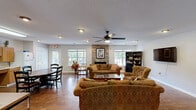 Located at 8825 Mont Carmel Ln in Charlotte, NC, this single-floor space offers a thoughtfully designed layout that balances comfort and functionality. The living room serves as the centerpiece of the space, highlighted by a striking stacked-stone fireplace that anchors the room with warmth and character. Rich hardwood-style flooring flows throughout, complementing the earthy tones of the upholstered seating. A ceiling fan with integrated lighting adds both practicality and comfort, while recessed lighting ensures a well-lit environment. A wall-mounted flat-screen TV, built-in bookshelf, and a dedicated workstation area with dual monitors make this living space as versatile as it is inviting. Natural light filters in through windows that offer views of the surrounding greenery. The kitchen is efficiently laid out with warm oak cabinetry extending from floor to ceiling, providing generous storage. Light-toned countertops offer ample prep space, and a double-basin stainless steel sink sits conveniently beneath the upper cabinets. A full-size refrigerator and microwave complete the kitchen's appliances. A dining area with a round wood table is positioned adjacent to the kitchen, creating a seamless flow between cooking and dining. The textured ceilings add a classic finishing touch throughout.