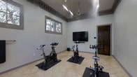 This dedicated spin room offers a purpose-built environment for indoor cycling enthusiasts. The space features multiple stationary spin bikes, each positioned on protective floor mats atop light-toned tile flooring, providing ample room between stations for a comfortable workout experience. A large wall-mounted flat-screen television serves as the focal point of the room, ideal for streaming cycling classes or entertainment during sessions. A ceiling fan with a sleek fixture ensures consistent air circulation throughout the space, keeping the environment cool during intense workouts. The room benefits from natural light filtering through a multi-pane window, offering a pleasant connection to the outdoors. Elevated ceilings with detailed crown molding trim lend an unexpected architectural refinement to the functional space, while the neutral gray walls contribute to a clean, focused atmosphere. A solid wood door provides a private entry point, reinforcing the room's designation as a standalone fitness area. The overall design strikes a balance between practicality and comfort, making it a well-appointed space for group or individual cycling training.