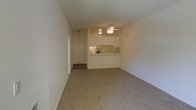 This single-floor residential unit offers a clean, functional layout designed for comfortable everyday living. The main living area is an open, carpeted space with neutral-toned walls and a textured ceiling, creating a bright and welcoming atmosphere. A ceiling fan with an integrated light fixture is centrally positioned, providing both air circulation and ambient lighting to the room.

The kitchen is situated at the far end of the living area, separated by a raised pass-through bar counter that adds a sense of openness while defining the two spaces. The kitchen features tile backsplash detailing and appears equipped with standard appliances and counter space. The pass-through design allows for easy interaction between the kitchen and living area, making the space feel connected and sociable.

A hallway visible from the main living area leads to additional spaces within the unit, including what appears to be a closet or utility door. The entryway features a darker flooring transition, distinguishing it from the carpeted living space.

Throughout the unit, the consistent use of light, neutral colors on the walls and flooring contributes to an airy, understated aesthetic. The layout is practical and efficient, making good use of the available floor space while maintaining a sense of openness and flow.