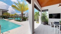 This single-story space is defined by its resort-inspired outdoor design, centered around a shimmering pool that serves as the focal point of the amenity area. The covered lanai showcases a warm wood-paneled ceiling and crisp white structural columns, creating a striking contrast that blends contemporary and tropical architectural elements. Large open sightlines from the covered area allow unobstructed views of the pool and surrounding landscape, enhancing the seamless indoor-outdoor connection.

The pool deck is finished in light-toned pavers, maintaining a clean and cohesive aesthetic throughout the space. Aqua-colored lounge chairs are positioned poolside beneath shading structures, offering dedicated areas for relaxation. Tall, mature palm trees and lush tropical plantings frame the perimeter, adding both privacy and a verdant, resort-like ambiance.

Oversized decorative planters with tropical foliage accent the covered lanai, reinforcing the botanical theme. The space benefits from abundant natural sunlight, with the open-air design allowing full exposure to the sky and surroundings. The overall atmosphere is bright, airy, and polished, reflecting a well-designed outdoor living environment suited to a warm, tropical climate.