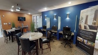 Located at 4001 W Northgate Dr in Irving, Texas, this single-story community clubhouse offers a well-appointed gathering space designed for both productivity and relaxation. The interior is characterized by a bold color palette, combining deep cobalt blue and warm terracotta orange accent walls that create a lively yet welcoming atmosphere throughout.

The business center area features multiple individual workstations, each equipped with a desktop computer and separated by partial privacy dividers mounted against the striking blue wall. Stylish hourglass-shaped wall sconces provide focused lighting at each station, while large multi-pane windows along the adjacent wall flood the space with natural light and offer views of the surrounding greenery. A bistro-height round table with leather chairs provides an additional spot for casual collaboration or meetings.

The lounge area is furnished with a set of sleek black leather armchairs arranged around a low coffee table, complemented by a wall-mounted flat-screen television and a colorful piece of abstract artwork. The open common area transitions seamlessly between zones, featuring a mix of wood-look and carpeted flooring, a brick accent column, a fireplace, and bright yellow seating that adds a cheerful contrast to the overall design scheme.