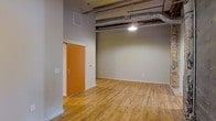 This single-floor space showcases a distinct industrial character defined by its high ceilings with exposed ductwork and ventilation systems running overhead. The warm-toned hardwood floors extend throughout, adding a natural contrast to the raw, utilitarian elements above. A pendant light fixture hangs from the ceiling, providing focused illumination and reinforcing the loft-inspired aesthetic. The walls are finished in a neutral gray tone, offering a clean, versatile backdrop suited to a variety of uses. A sliding door with a warm wood finish provides access to an adjoining area, contributing both functional and visual interest. The open layout creates a flexible environment with ample room for various configurations. Ventilation components are left intentionally visible, lending the space an authentic, converted-warehouse feel. The overall ambiance balances rugged industrial design with practical livability, making it a distinctive space that appeals to those who appreciate character-driven architecture combined with open, adaptable interiors.