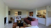 This single-story space showcases an open-concept living area that flows seamlessly into the kitchen, creating a connected and functional layout. The living room is furnished with a mauve sofa and a white armchair, both accented with patterned throw pillows in warm, earthy tones. A dark rectangular coffee table anchors the seating area, complemented by a patterned area rug that ties the color palette together. Wall art in rich amber and brown hues adds visual interest to the neutral-painted walls, while a floor lamp provides warm ambient lighting. The textured ceiling adds a subtle architectural detail overhead. The kitchen is visible through a pass-through opening, featuring white cabinetry and countertops with light-colored finishes, contributing to the overall brightness of the space. The open sightlines between the living and kitchen areas enhance the sense of spaciousness. The overall ambiance is warm and inviting, with a cohesive color scheme of mauve, brown, and white tones creating a comfortable and well-coordinated interior.