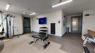 This fitness center offers a comprehensive workout environment designed to support a wide range of exercise routines. The space is laid out across a single floor, with durable speckled flooring that transitions between carpeted and hard-surface zones to accommodate different types of training activities.

The main workout area is furnished with a variety of cardio equipment, including a True treadmill and multiple stationary bikes, positioned near windows that allow natural light to fill the room. A full-length mirrored wall spans one side of the space, visually expanding the room and providing functional feedback during exercise. Ceiling fans are installed throughout, ensuring comfortable airflow during workouts.

A dedicated free weights section features a multi-tier dumbbell rack stocked with a complete set of dumbbells, paired with an adjustable workout bench for strength training. A wall-mounted flat-screen television displaying the Binary fitness platform is positioned above the weight rack, enabling guided workout sessions. A rolled exercise mat is conveniently stored nearby.

Additional equipment includes a cable resistance machine, TRX suspension trainer, and a ball rack holding stability balls in varying sizes. Cubbies for personal storage are available along one wall, and the space benefits from both recessed and strip lighting to maintain bright, consistent illumination throughout.