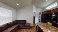 This single-floor unit offers a well-organized layout that efficiently combines living, cooking, sleeping, and utility functions. The open-concept living and kitchen area forms the heart of the space, anchored by warm wood-look hardwood flooring that extends throughout. Vaulted ceilings in the living area lend a sense of added height and openness, while large windows fitted with horizontal blinds allow abundant natural light to fill the room. A generously sized sectional sofa fits comfortably within the living space, which also benefits from recessed lighting overhead.

The kitchen is outfitted with rich, dark wood cabinetry, granite countertops, and a full suite of black appliances including a refrigerator, electric range, over-the-range microwave, and dishwasher. A kitchen island with a granite surface provides additional workspace. Elevated transom windows above the cabinetry introduce extra natural light. A stacked washer and dryer unit is conveniently tucked behind a paneled door adjacent to the kitchen.

The bedroom features high clerestory windows, a wall-mounted mini-split system for climate control, a closet, and a wooden dresser. The full bathroom includes dark wood vanity cabinetry, granite countertops, a glass-enclosed shower-tub combination, a window for natural light, and wood-look flooring consistent with the rest of the unit.
